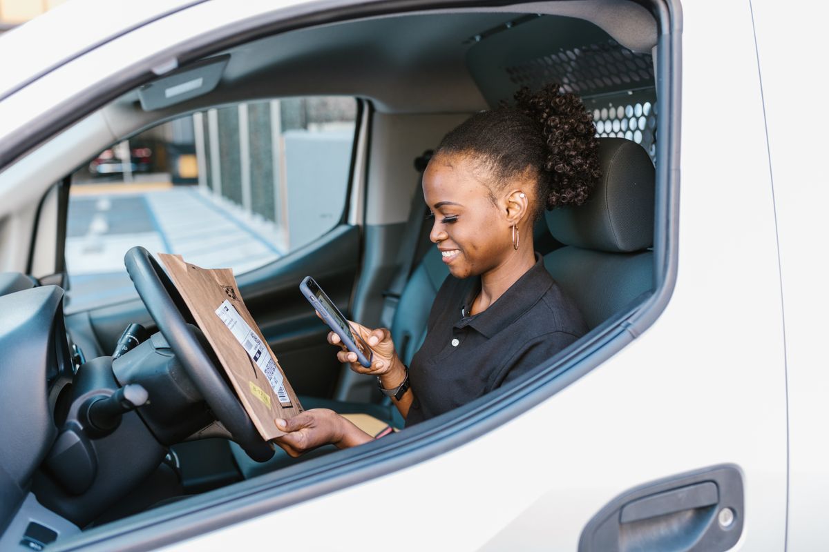 Lächelnde Kurierfahrerin mit Paket und Smartphone am Steuer ihres Lieferwagens